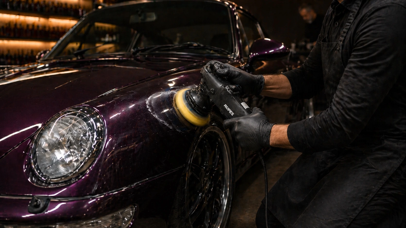 Detailer in black gloves operating a Rupes random-orbital polisher on the front fender of an aubergine Porsche 993 Turbo, under warm studio lighting